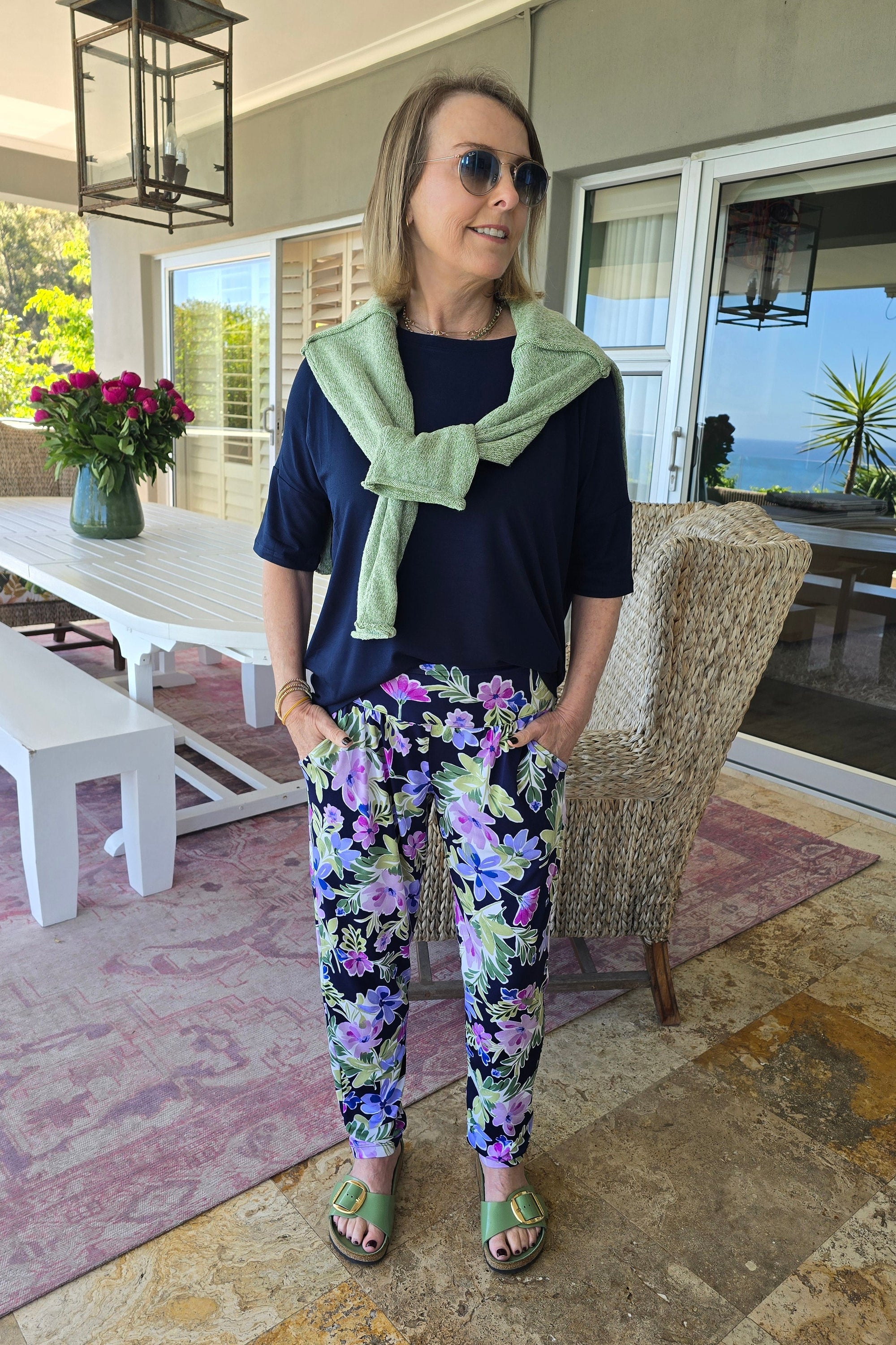 Woman standing on a patio wearing a dark top and floral pants with a green cardigan.